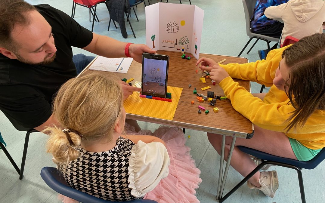 A man and two girls looking at a lego scene through a tablet device